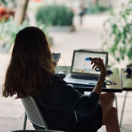 Frau sitz auf einem Outdoor-Stuhl vor einem MacBook und redet in ein iPhone