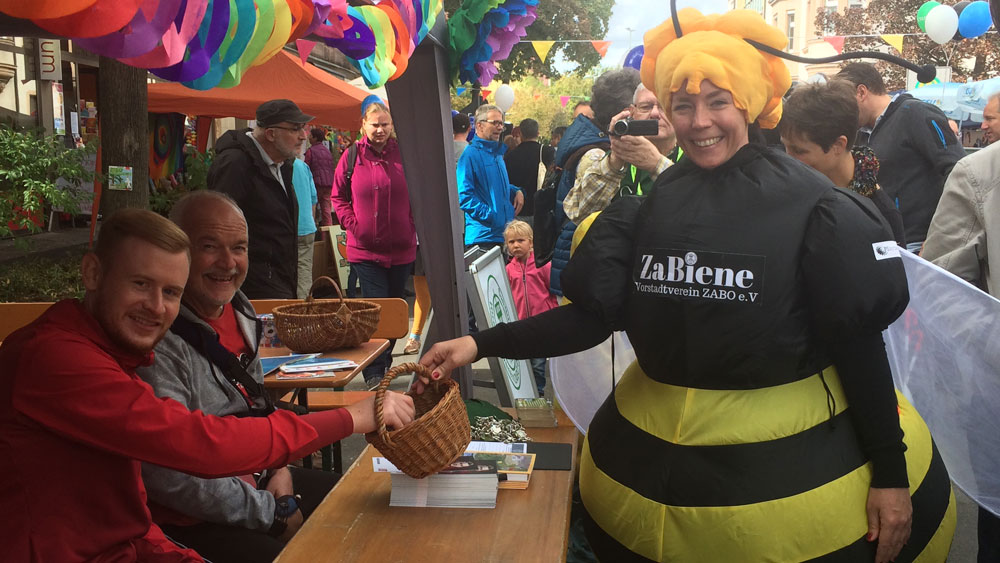 Eine Frau in Bienenkostüm reicht Club-Profi Sebastian Kerk einen Korb am Infostand des Vorstadtvereins ZABO e.V