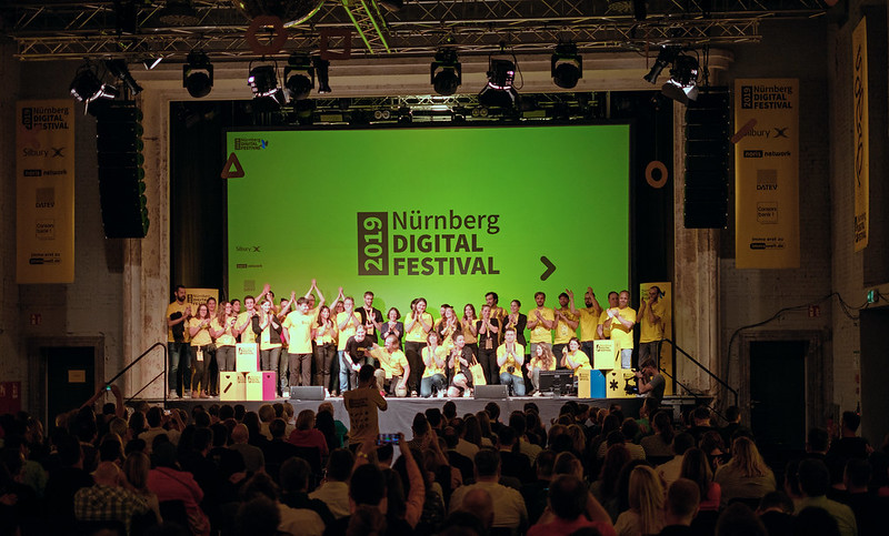 Viele Menschen in gelben T-Shirts stehen klatschend auf einer Bühne und bekommen Applaus. Im Hintergrund ist auf DEr Bühnenleinwand "Nürnberg Digital Festival 2019 " zu lesen.