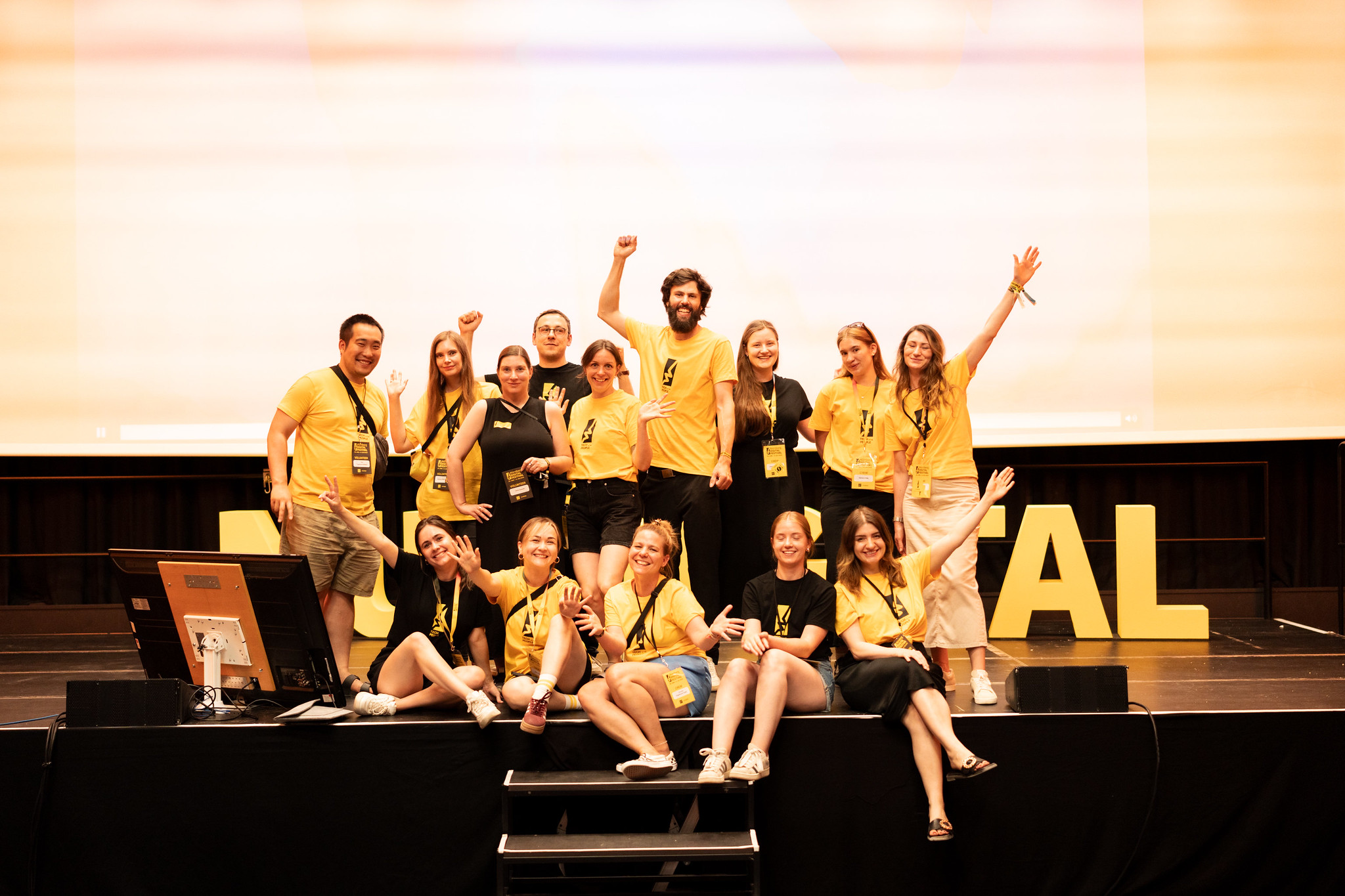 Group of young people in yellow and black T-shirts smiling and cheering on stage in front of large lettering.