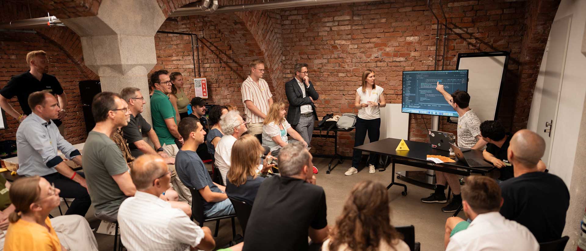 Gruppe von Menschen sitzt in Ziegelraum und verfolgt Präsentation, während Person auf Bildschirm mit Code zeigt.