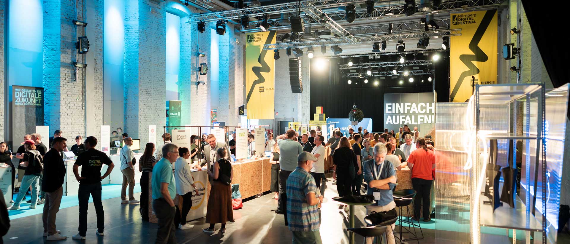 Menschen schlendern durch helle Ausstellungshalle mit Infoständen, gelben Festivalbannern und leuchtender Aufschrift „EINFACH AUFALLEN!“ im Hintergrund.