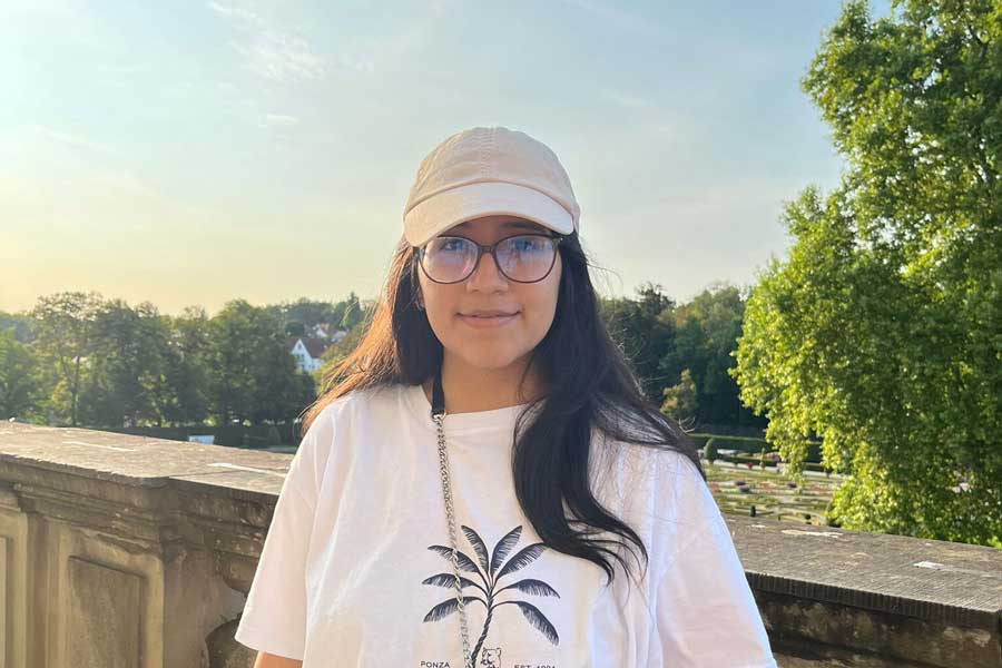 Junge Frau mit langem dunklem Haar, Brille und beigefarbener Kappe steht draußen vor einer steinernen Brüstung. Sie trägt ein weißes T-Shirt mit schwarzem Palmenmotiv und blickt in die Kamera. Im Hintergrund Bäume, Wiesen und Gebäude bei Sonnenuntergang.