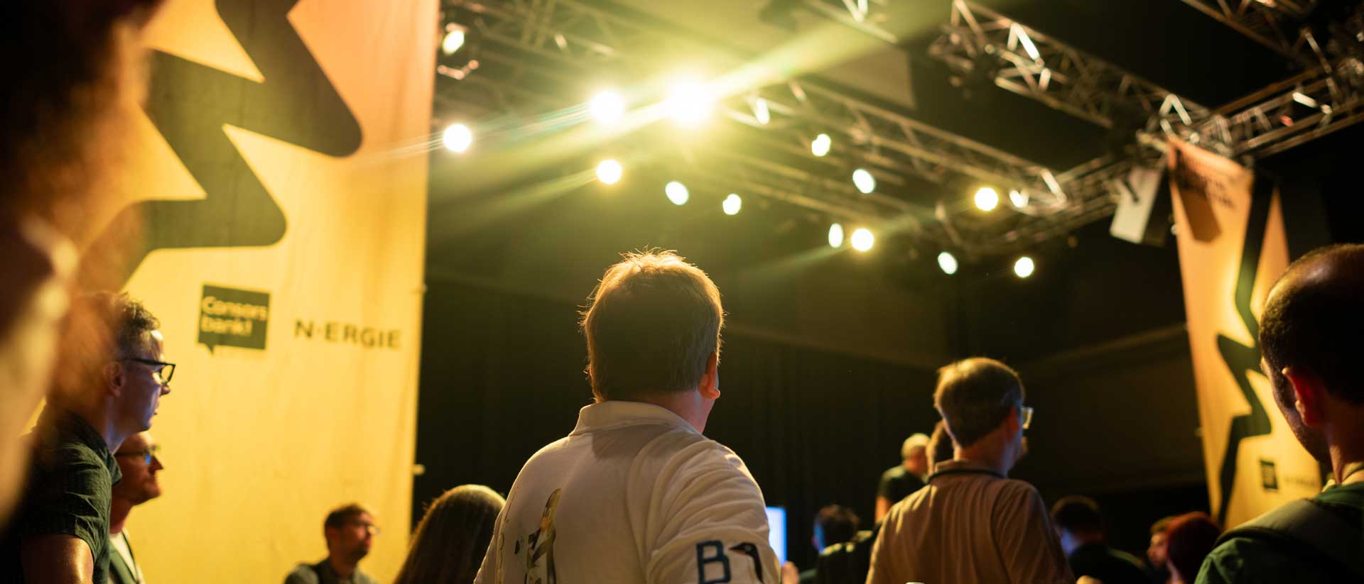 Menschen stehen in einem beleuchteten Veranstaltungsraum vor einer Bühne. Große gelbe Banner mit Firmenlogos hängen seitlich. Eine Wasserflasche und ein kleines Schild stehen auf einem Tisch im Vordergrund.