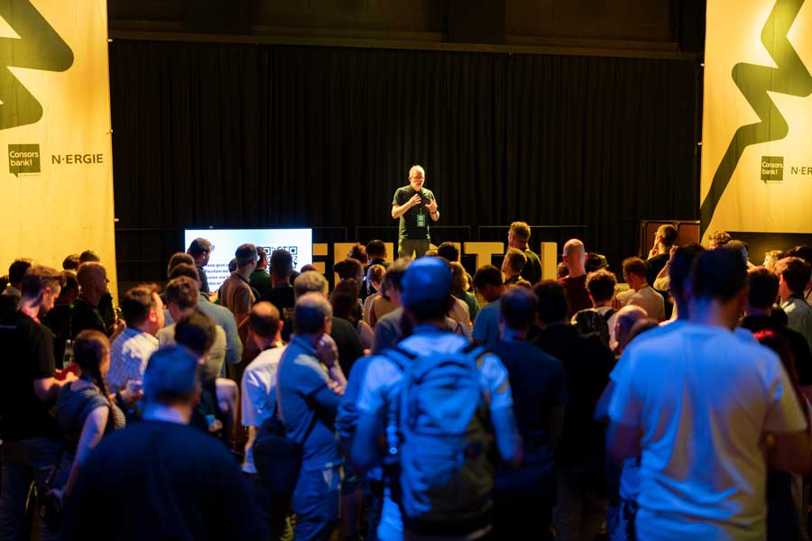 Mann spricht auf Bühne vor dicht gedrängtem Publikum, links daneben Bildschirm mit QR-Code, schwarze Vorhänge im Hintergrund.