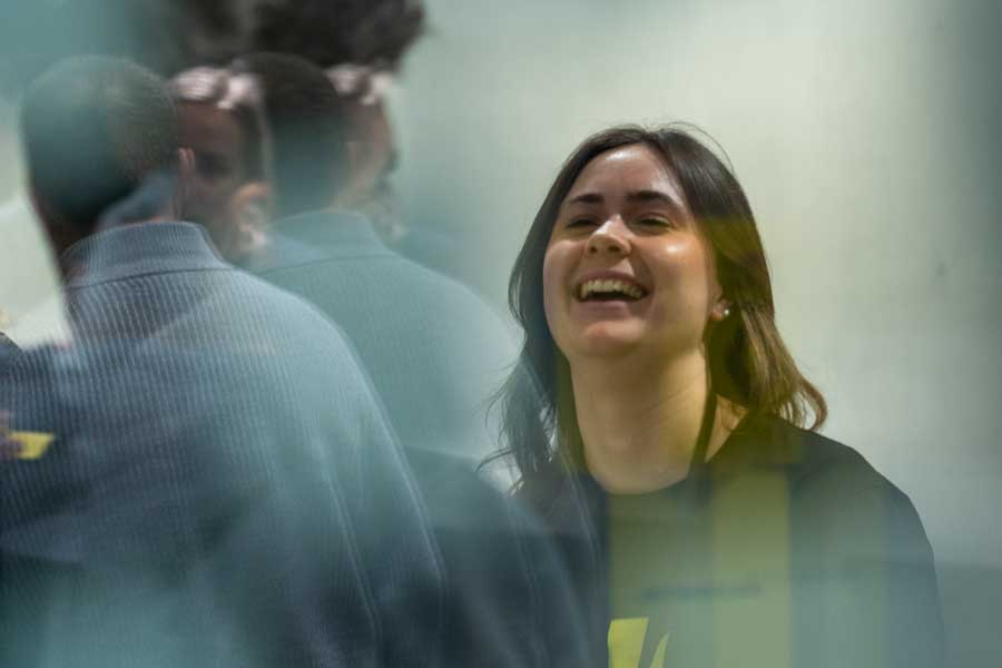 Lachende Frau mit dunklen Haaren steht in einem Raum, im Vordergrund spiegeln sich mehrere Personen.