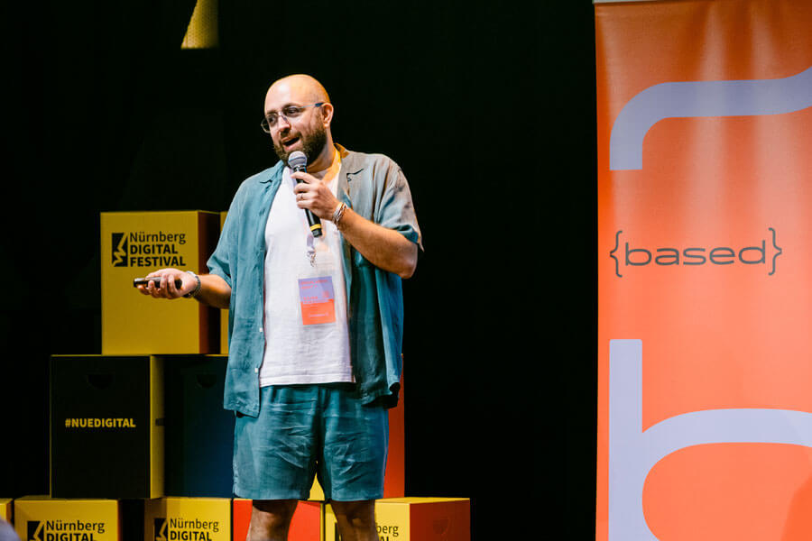 Mann mit Mikrofon steht auf Bühne vor gelben Boxen mit Nürnberg Digital Festival Logo und einem orangefarbenen Banner mit der Aufschrift „based“.