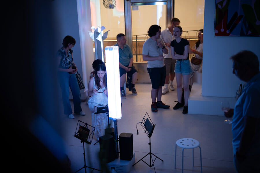 Mehrere Personen in abgedunkeltem Raum, eine Frau sitzt vor heller LED-Leuchte mit Technikgeräten, andere stehen im Hintergrund.