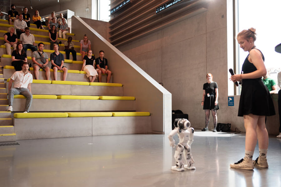 Frau mit Mikrofon steht neben einem kleinen humanoiden Roboter, mehrere Personen sitzen auf gelber Tribüne im Hintergrund.Frau mit Mikrofon steht neben einem kleinen humanoiden Roboter, mehrere Personen sitzen auf gelber Tribüne im Hintergrund.
