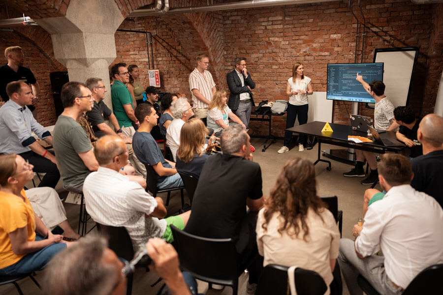 Gruppe sitzt in Kellerraum mit Ziegelwänden, zwei Personen zeigen auf Code auf einem Bildschirm, gelbes Nürnberg Digital Festival Schild auf dem Tisch.Gruppe sitzt im Raum mit Ziegelwänden, zwei Personen zeigen auf Code auf einem Bildschirm, gelbes Nürnberg Digital Festival Schild auf dem Tisch.