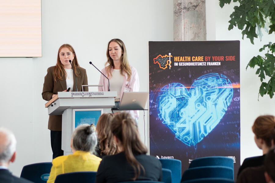 Zwei Frauen stehen an einem Rednerpult mit Mikrofon, rechts daneben Banner mit Herzgrafik und Text „Health Care by Your Side“.
