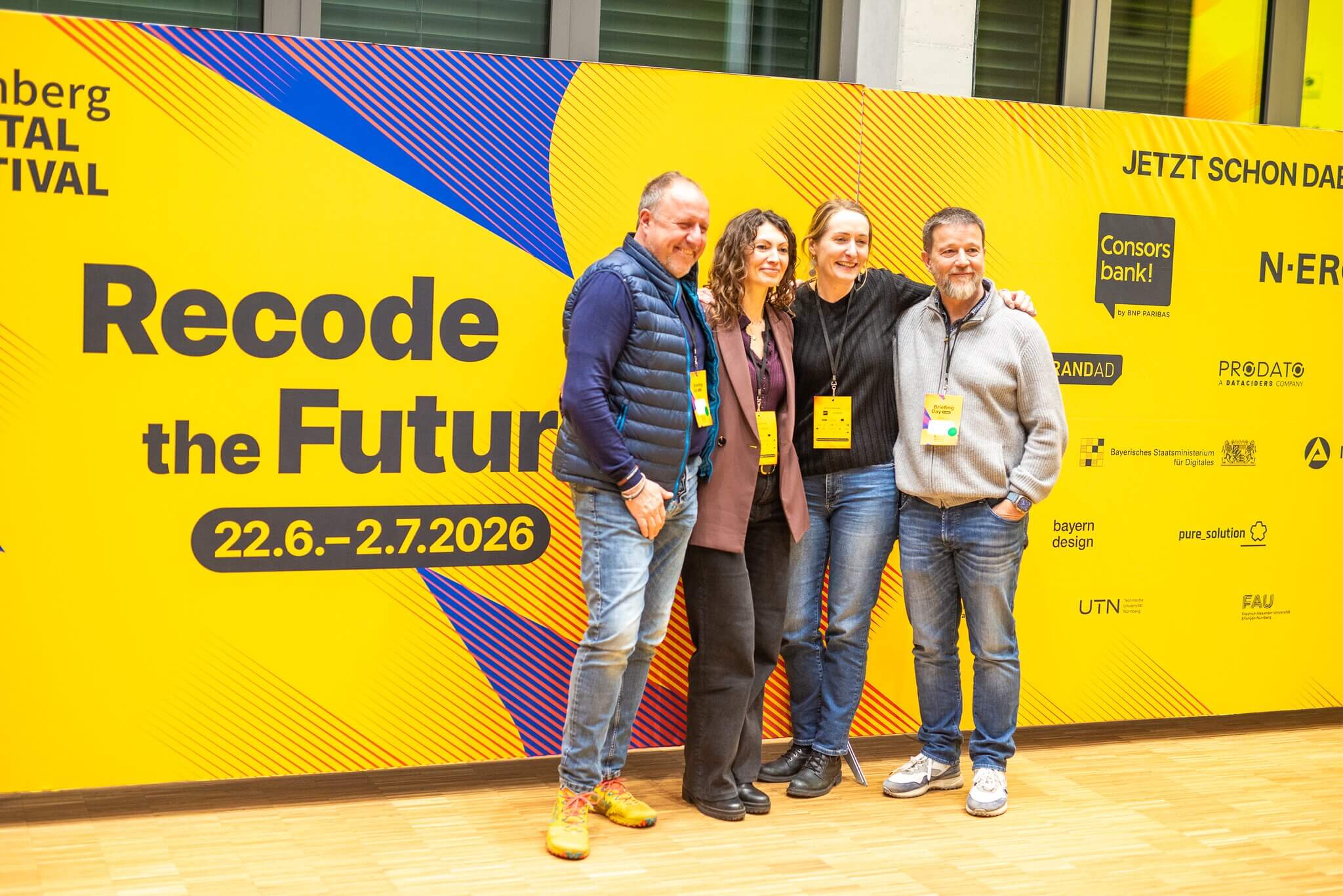 Four people smiling in front of a yellow wall with the black text “Recode the Future 22.6.–2.7.2026” and multiple sponsor logos.