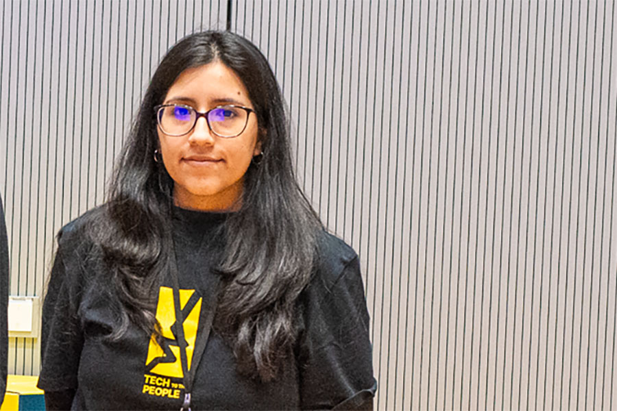 Junge Frau mit langem dunklem Haar, Brille und beigefarbener Kappe steht draußen vor einer steinernen Brüstung. Sie trägt ein weißes T-Shirt mit schwarzem Palmenmotiv und blickt in die Kamera. Im Hintergrund Bäume, Wiesen und Gebäude bei Sonnenuntergang.