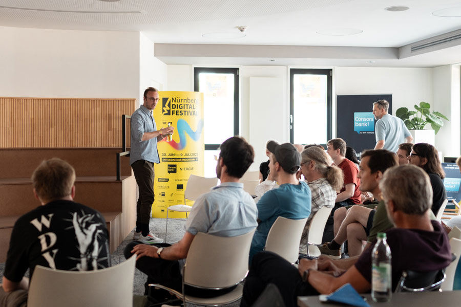 Man presenting in front of audience next to yellow Nürnberg Digital Festival stele in modern conference room.