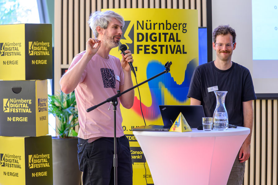 Two men at standing table with microphone and laptop in front of Nürnberg Digital Festival stele.