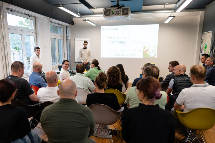 Men moderate interactive session in front of audience, presentation slide reads: “(First) name”, “Organization”, “My worst process experience”.