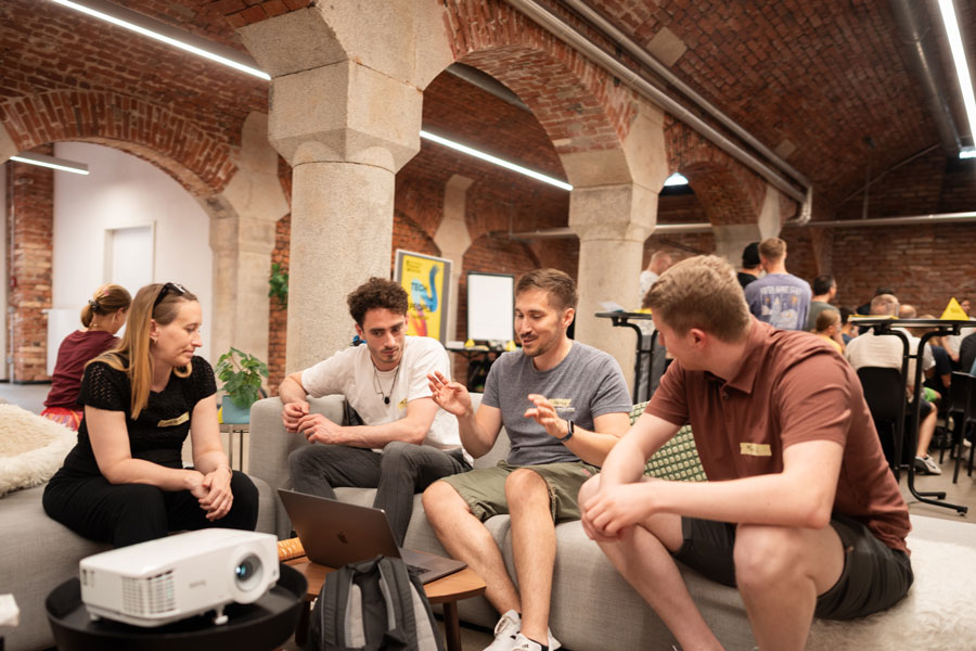Four people sit in a group on a sofa under brick arches, discussing in front of a laptop.