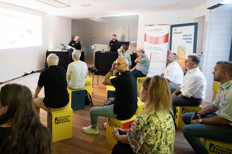 Group seated on yellow stools with Nürnberg Digital Festival logo watching a presentation with technical graphics.