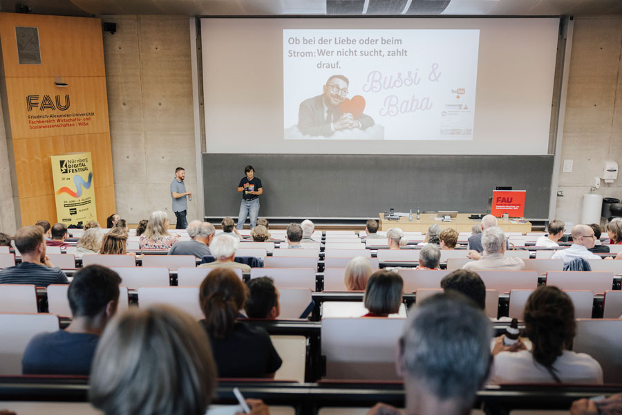 A woman and a man speak on stage in lecture hall, projected slide with text and image in background; audience seated in rows.