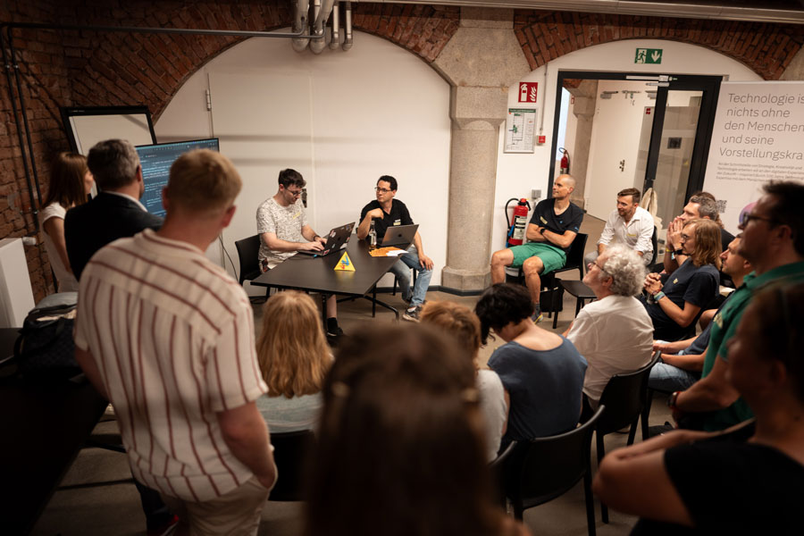 Group listening to two men at table with laptops in brick-walled room; flipchart and projection in background.