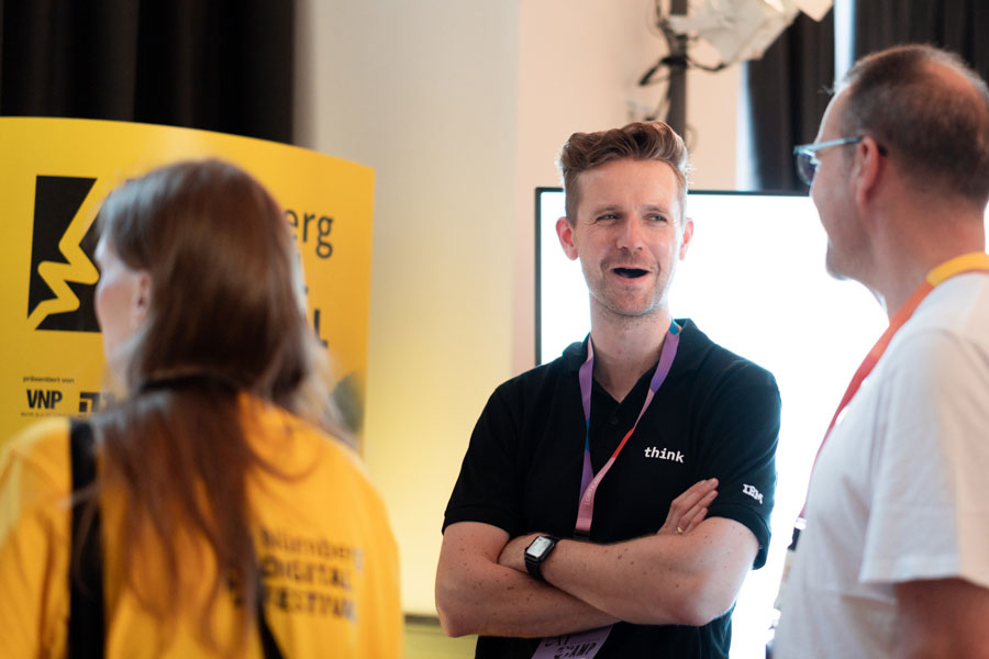 Drei Personen im Gespräch vor gelber Stele mit Text und Symbol, Mann in schwarzem Shirt mit „think“-Aufdruck.