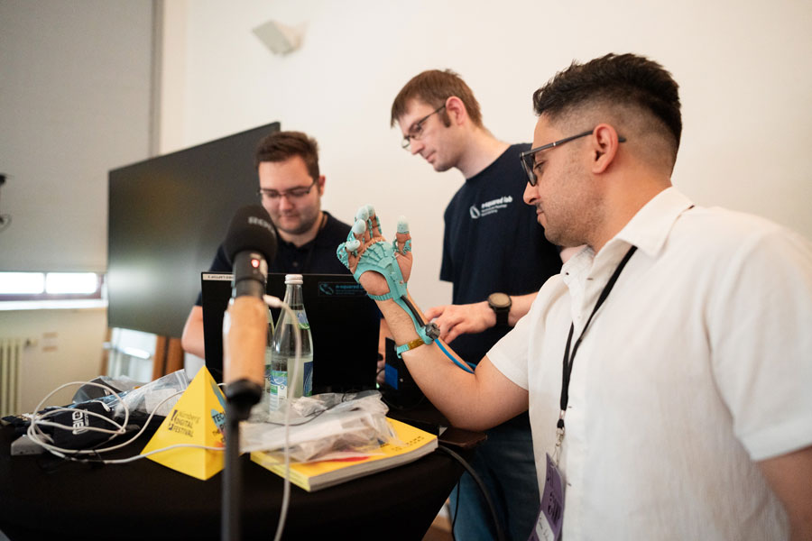 Mann zeigt blaues robotisches Handmodell, zwei weitere Männer arbeiten an Laptops im Hintergrund.