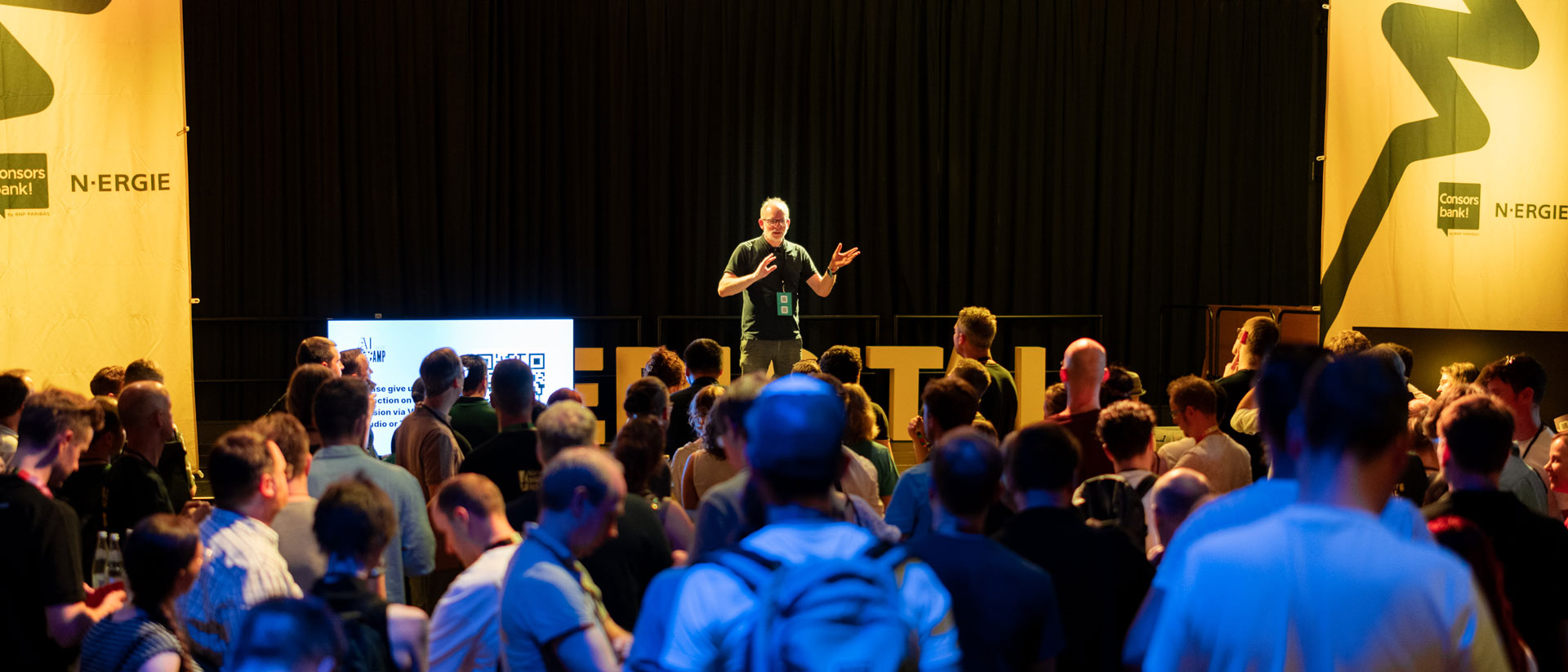 Sprecher steht auf Bühne vor Publikum, rechts und links gelbe Banner mit nuedigital Logo und Sponsor-Logos.