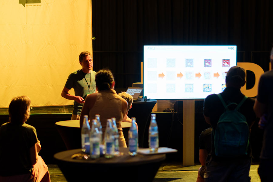 Mann präsentiert vor Bildschirm mit Pfeilgrafik, Zuhörer sitzen oder stehen im Halbkreis.