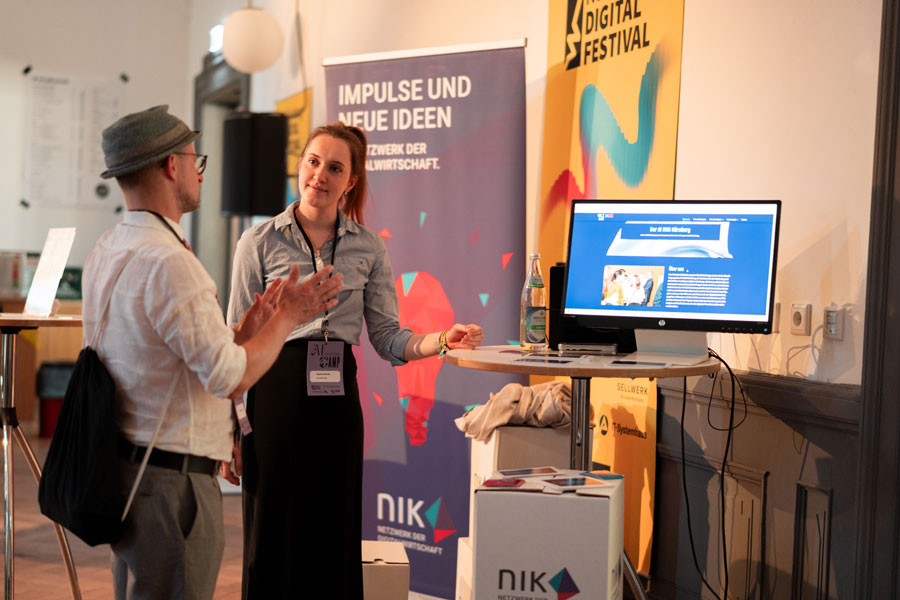Zwei Personen im Gespräch an Infostand mit Monitor, Roll-up mit Text „Impulse und neue Ideen“ im Hintergrund.