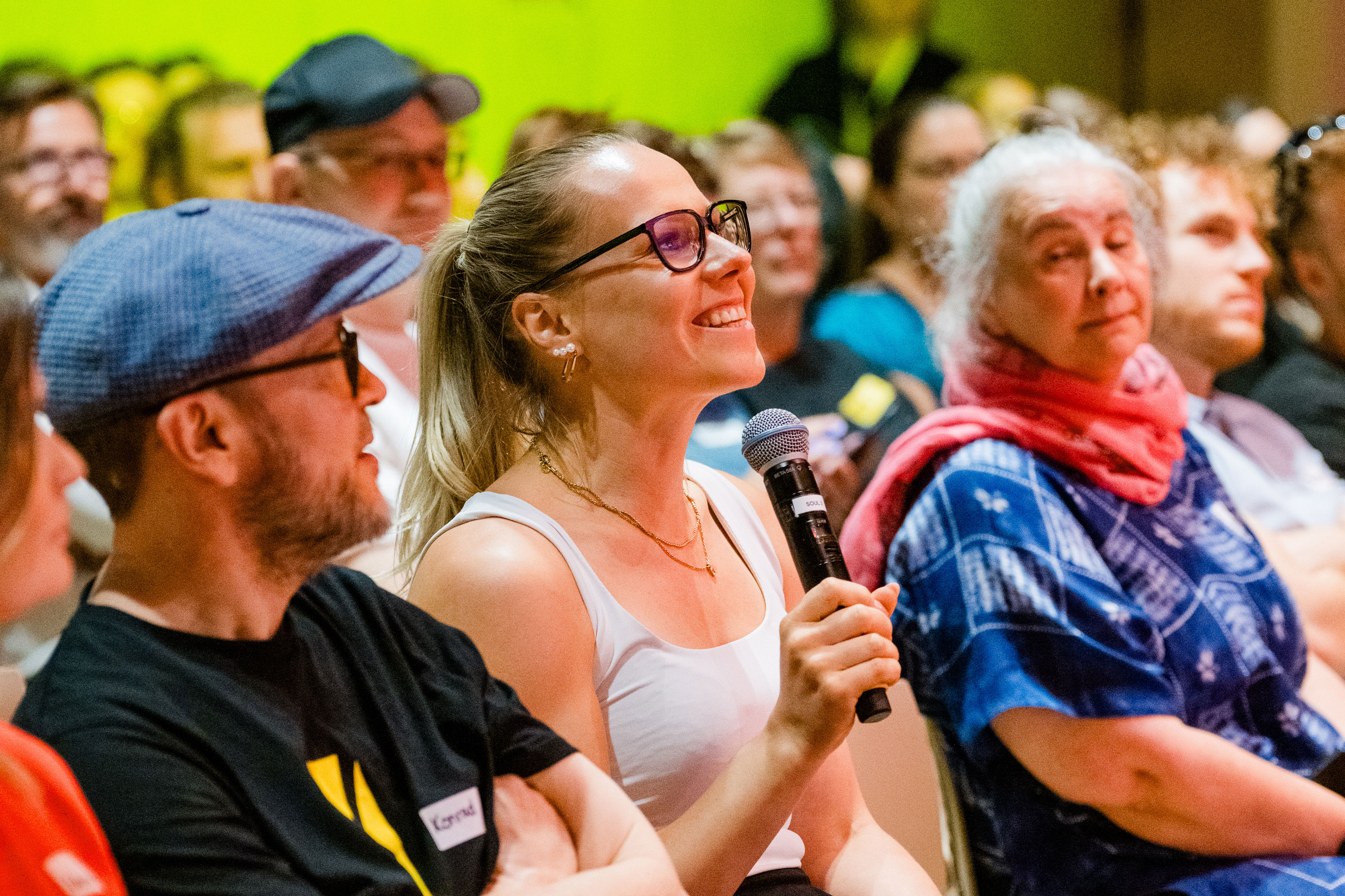 Lächelnde Frau mit Mikrofon sitzt in Publikum, spricht oder stellt Frage, umgeben von anderen Zuschauer:innen.