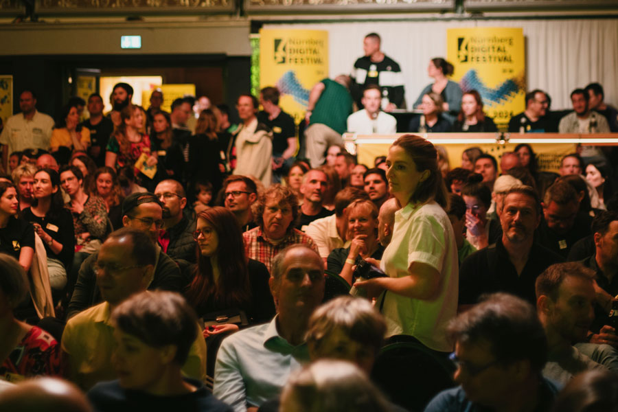 Großer Veranstaltungsraum mit dicht gedrängtem Publikum, Personen sitzen und stehen, Banner mit „Nürnberg Digital Festival“ im Hintergrund.