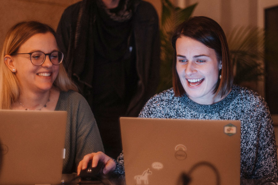Zwei Frauen mit Laptops, rechts vorn lachend; links mit Brille schaut zu, Person dahinter