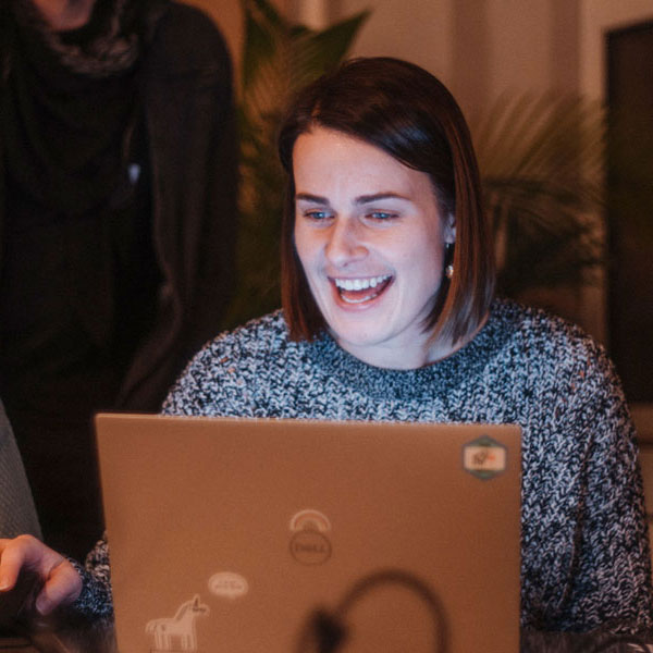 Lächelnde Frau sitzt vor Laptop, schaut auf den Bildschirm, weitere Person im Hintergrund.