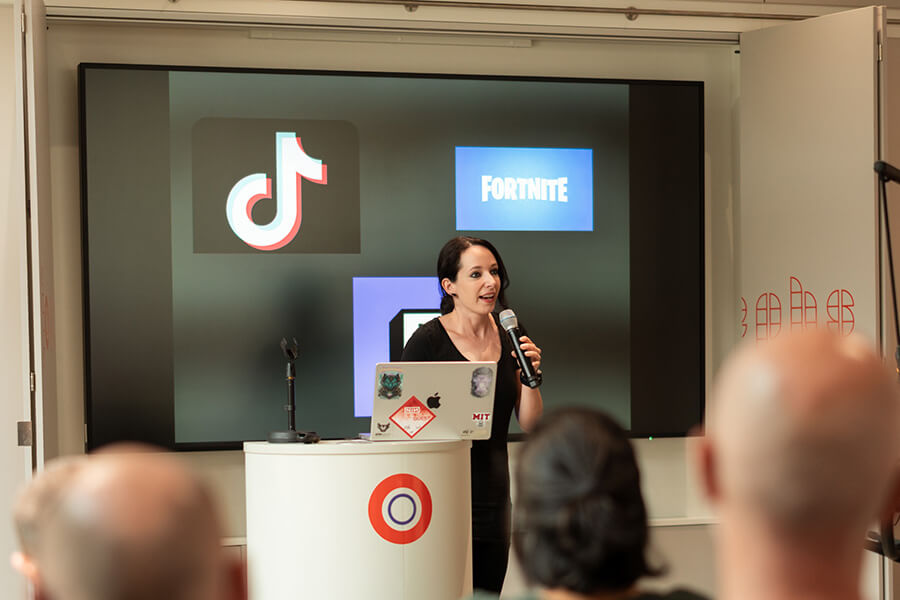 Frau mit Mikrofon steht rechts neben Podium mit Laptop; hinter ihr Logos TikTok und Fortnite.