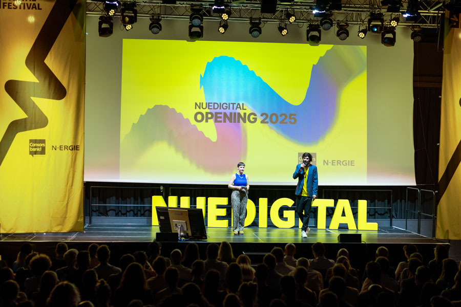 Bühne mit zwei Personen vor Leinwand „NUEDIGITAL OPENING 2025“, Publikum im Vordergrund