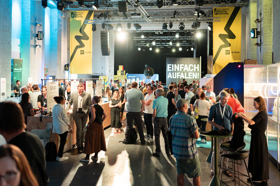 Große Halle mit Messeständen und vielen Menschen, Bühne hinten mit Schrift „EINFACH AUFFALLEN!“