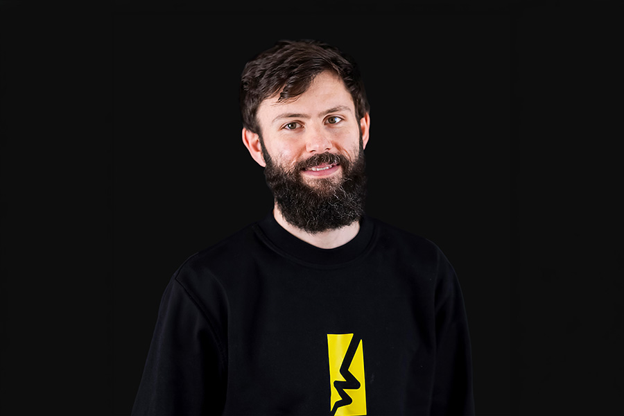Bearded man with microphone speaking to a group, yellow background.