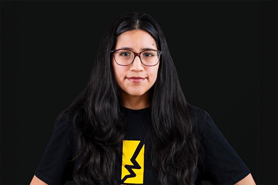 A young woman with long dark hair, glasses, and a beige cap is standing outside by a stone railing. She’s wearing a white T-shirt with a black palm tree design and looking straight at the camera. Behind her, there are trees, grassy fields, and some buildings lit by the sunset.