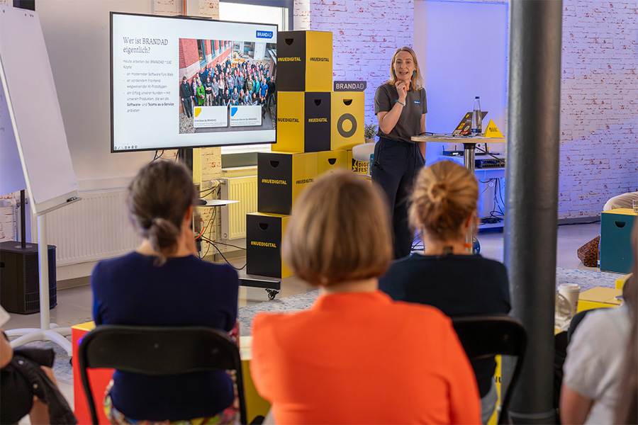 Frau präsentiert vor Bildschirm „Wer ist BRANDAD“, Publikum sitzt davor, gestapelte Kisten