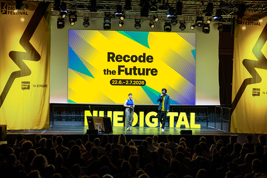 Bühne mit zwei Personen vor Leinwand „NUEDIGITAL OPENING 2025“, Publikum im Vordergrund