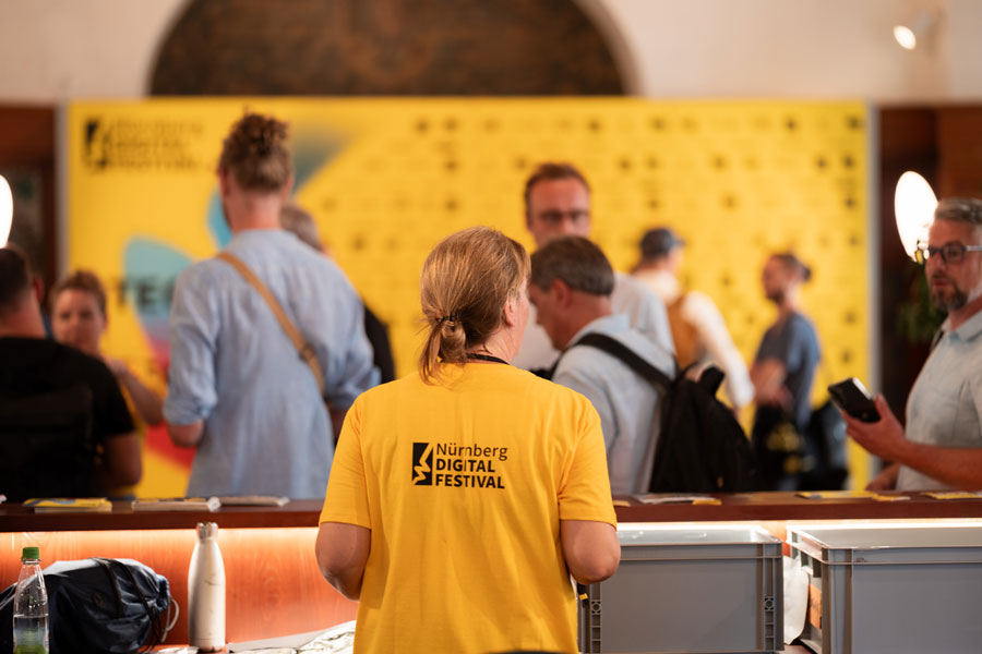 Person mit gelbem T-Shirt steht am Tresen, dahinter Besucher; Text „Nürnberg Digital Festival“
