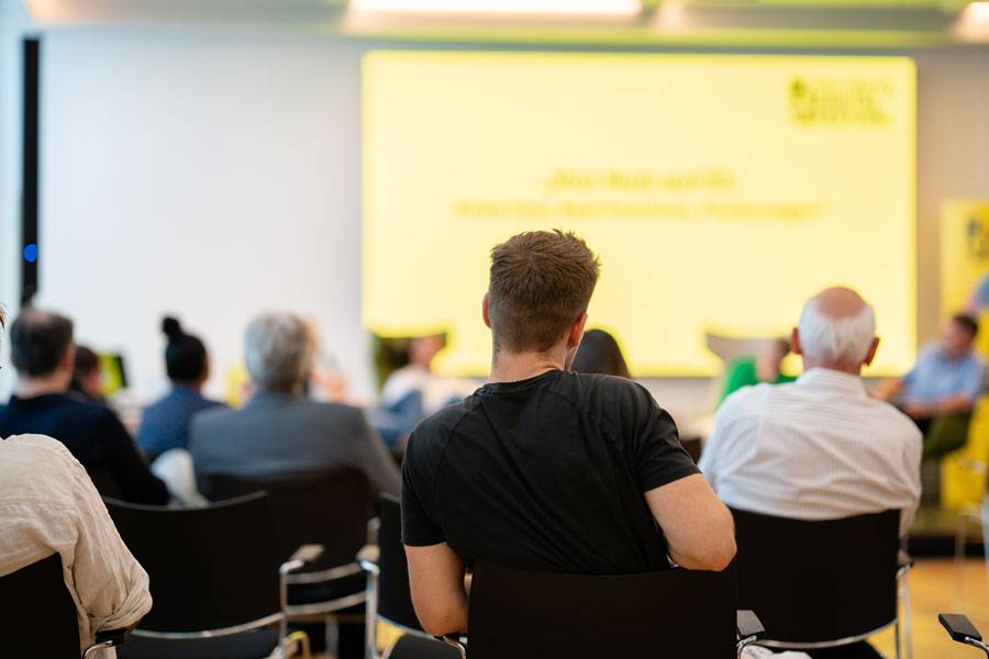 Publikum sitzt vor gelber Präsentationsfläche in hellem Veranstaltungsraum.