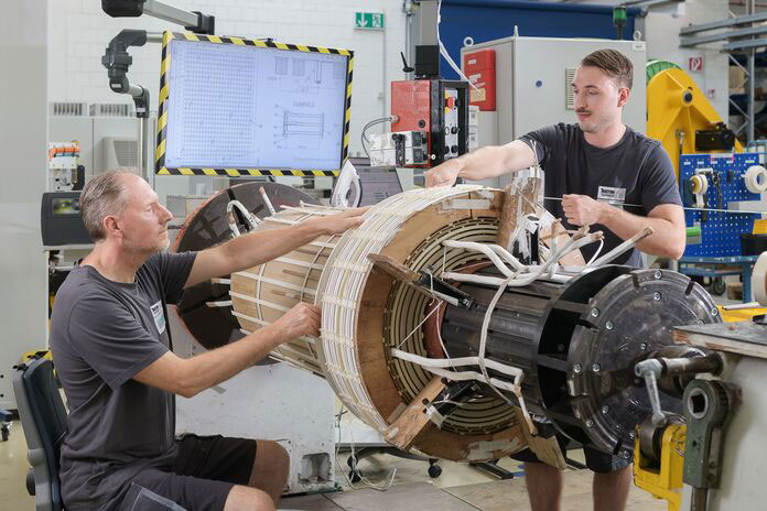 Zwei Männer arbeiten in Werkstatt an großer technischer Spule mit Kabeln.