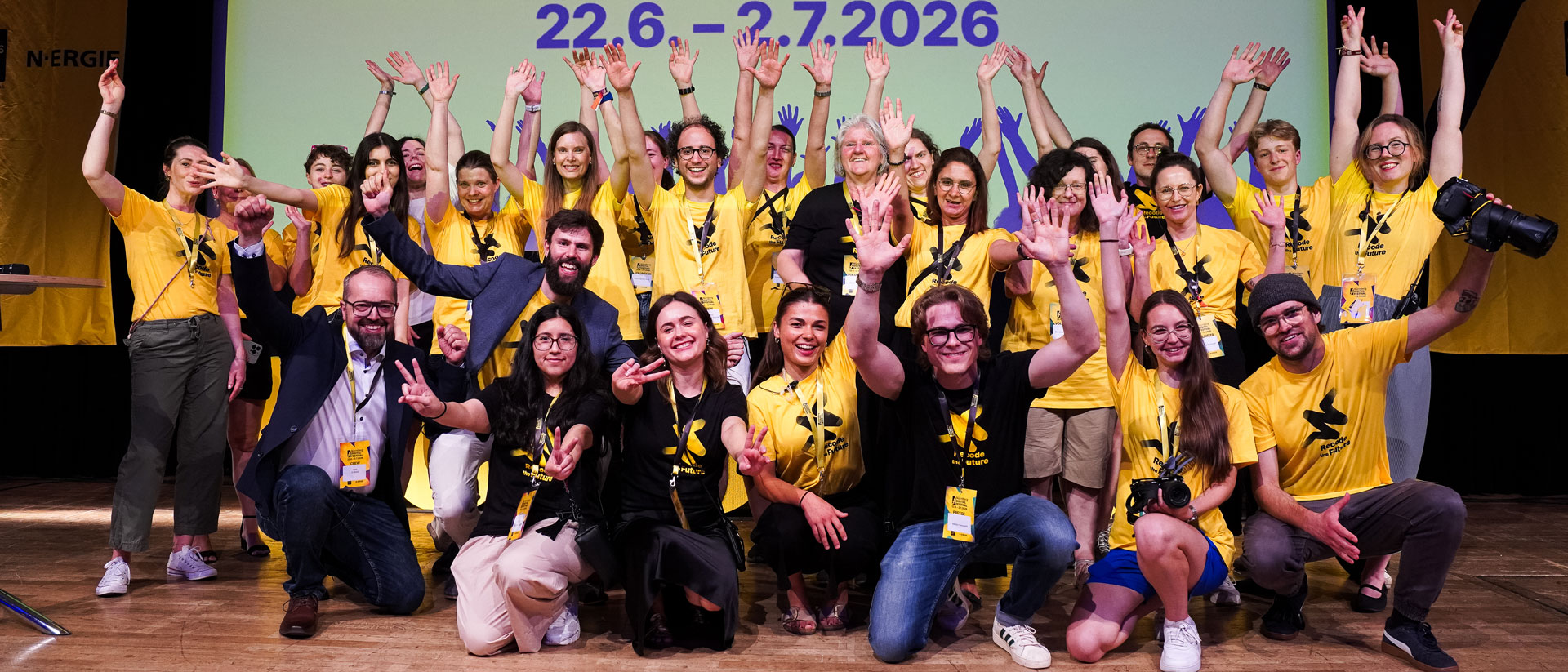 Große Gruppe in gelben Shirts winkt vor dem Datum „22.6.–2.7.2026“.