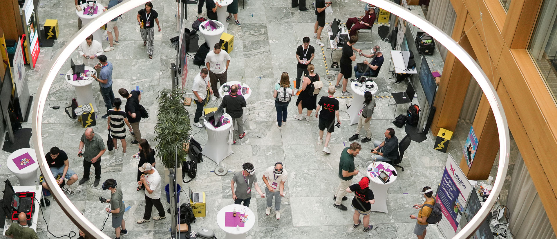 Blick von oben auf viele Personen an VR-Stationen und Stehtischen in einer Halle.