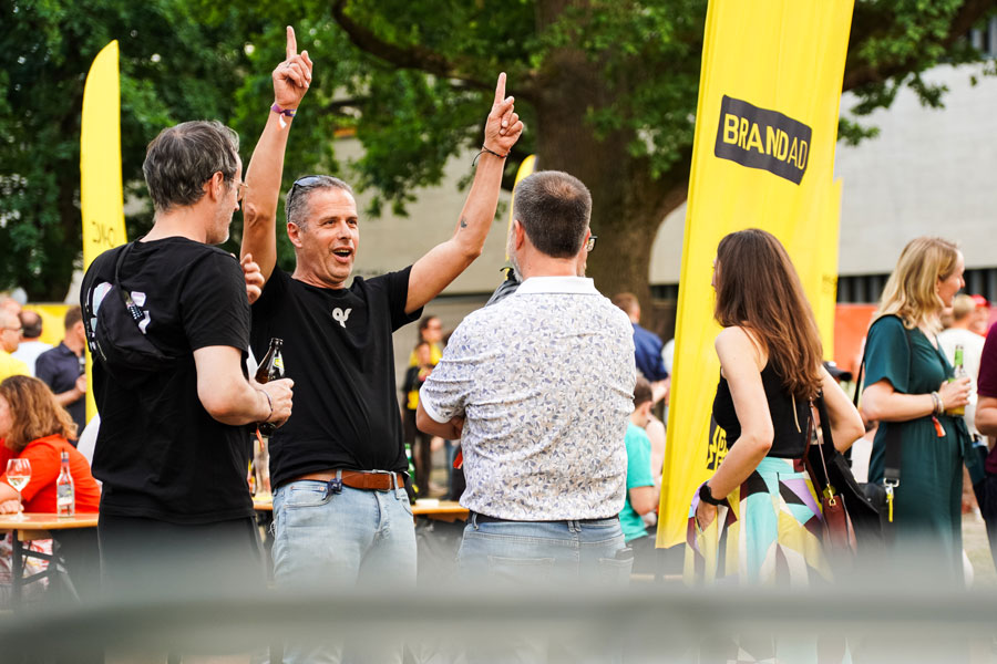 Mann hebt beide Arme zwischen zwei Männern; gelbes Banner trägt den Text „BRANDAD“.