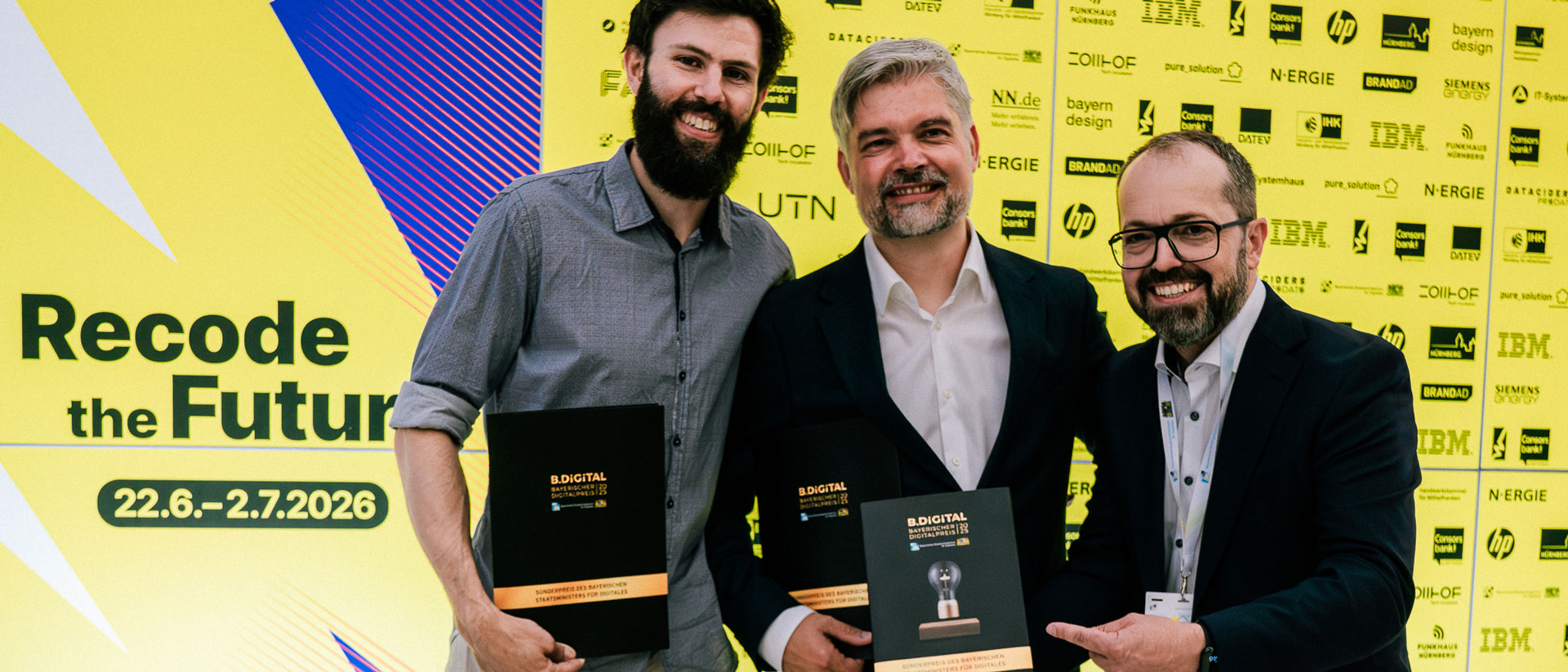 Drei lächelnde Männer stehen vor einer gelben Festival- und Sponsorenwand. Zwei von ihnen halten Urkunden und eine Trophäe des Bayerischen Digitalpreises B.DiGiTAL. Links ist das Festivalmotto „Recode the Future“ mit dem Zeitraum 22. Juni bis 2. Juli 2026 zu sehen.