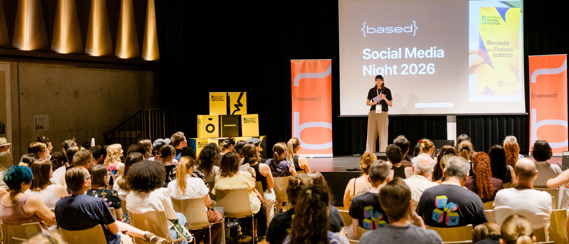 Eine Frau steht auf einer Bühne vor einer großen Leinwand mit der Aufschrift „Social Media Night 2026“. Vor ihr sitzt ein voll besetztes Publikum. Neben der Bühne stehen orangefarbene Banner und gelbe Festivalwürfel des Nürnberg Digital Festivals.