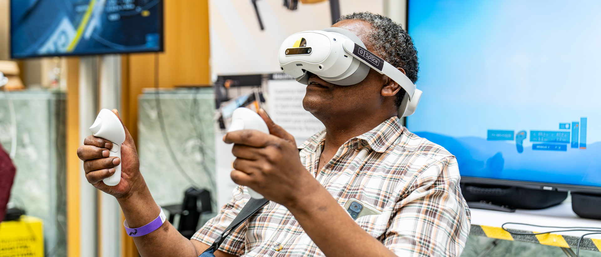 Ein Besucher trägt eine Virtual-Reality-Brille und hält zwei Controller in den Händen. Im Hintergrund sind Bildschirme und ein Ausstellungsstand des XR Day zu sehen.