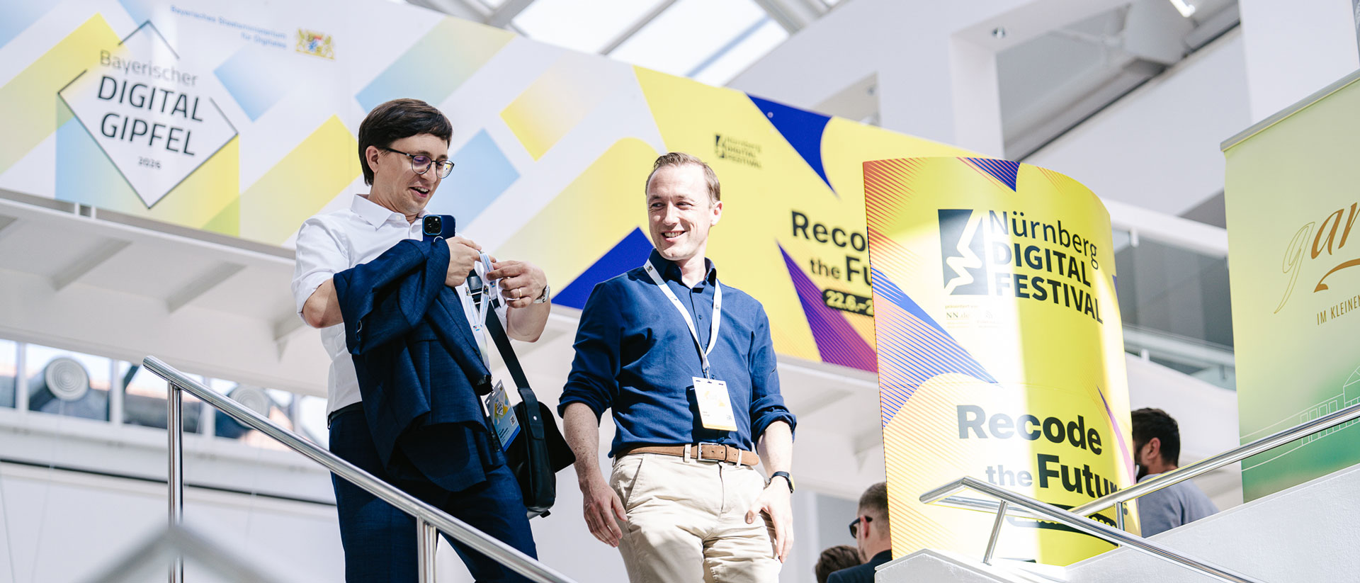 Zwei Männer mit Namensschildern unterhalten sich im hellen Foyer des Bayerischen Digitalgipfels. Neben ihnen stehen gelbe Aufsteller des Nürnberg Digital Festivals mit dem Motto „Recode the Future“.